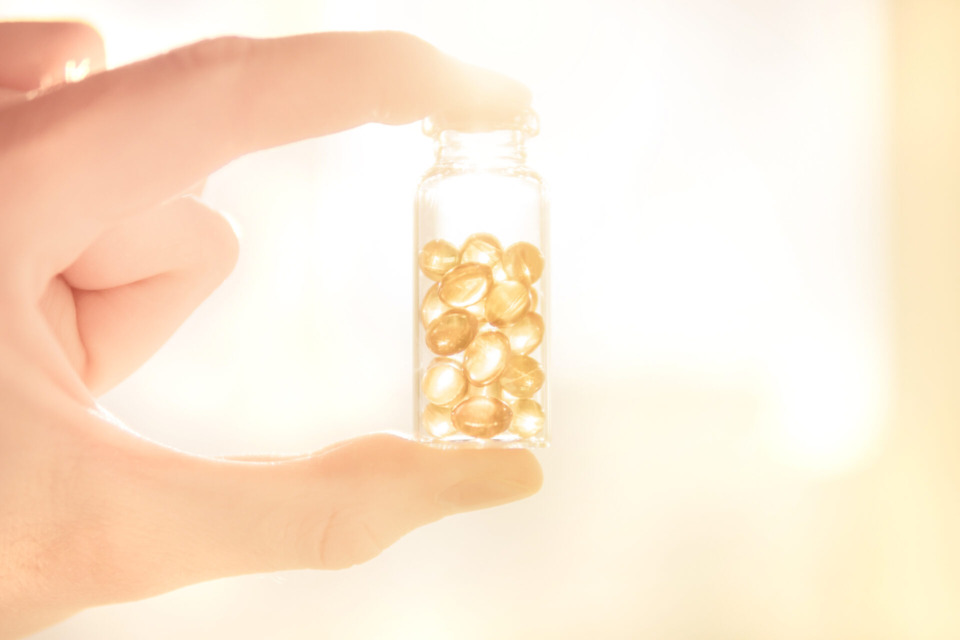 A hand holds a small glass jar filled with yellow capsules against a bright, light-filled background.