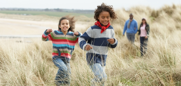 Two children run joyfully through tall grass in a sandy, beachside landscape, while two adults walk behind them, partially visible in casual clothing.