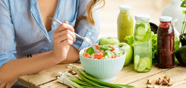 A person eats a salad with a fork at a wooden table, surrounded by fresh vegetables and colorful juice bottles, while wearing a blue-striped shirt.