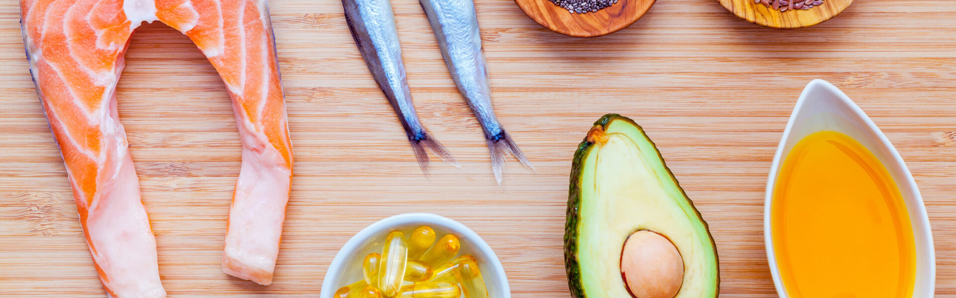 Salmon steak, small fish, avocado, omega-3 capsules, and bowls of seeds are arranged on a wooden surface, creating a display of healthy food items.