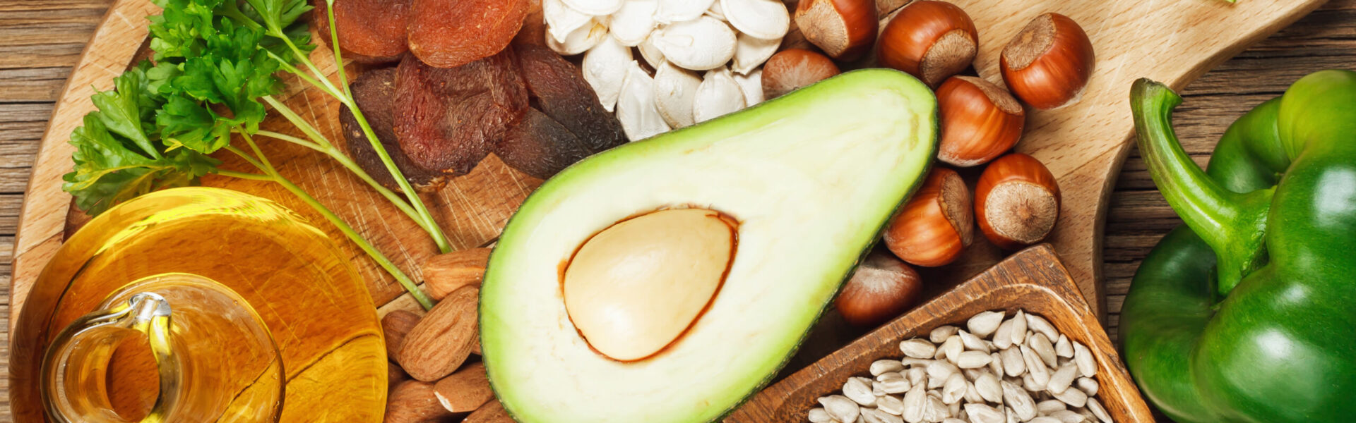 An avocado half rests on a wooden board surrounded by parsley, oil, dried apricots, pumpkin seeds, hazelnuts, almonds, sunflower seeds, and a green bell pepper.