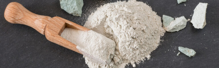 Wooden scoop holds fine white powder, resting on a dark surface with scattered chunks of light green rock nearby.