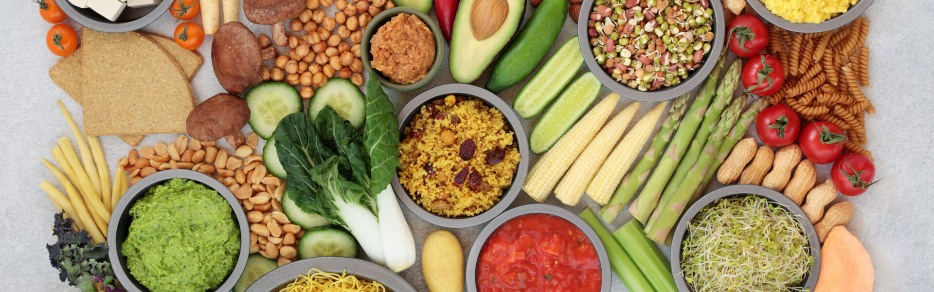 An assortment of colorful plant-based foods, featuring bowls of dips, vegetables, fruits, nuts, grains, and sprouts, arranged neatly on a light surface.