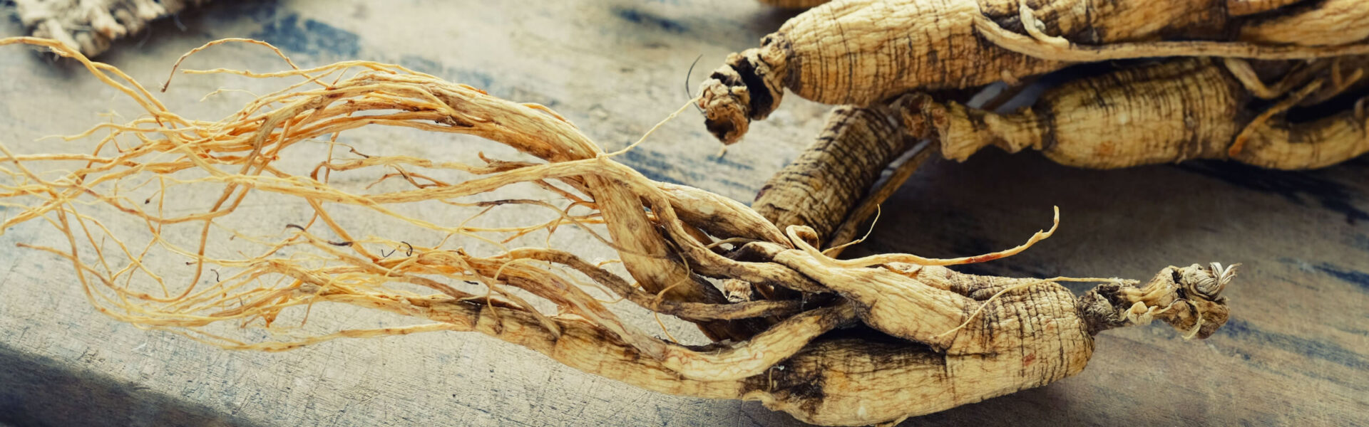 Dried ginseng roots lie on a wooden surface, their twisting forms and fine tendrils creating intricate patterns against the textured backdrop.