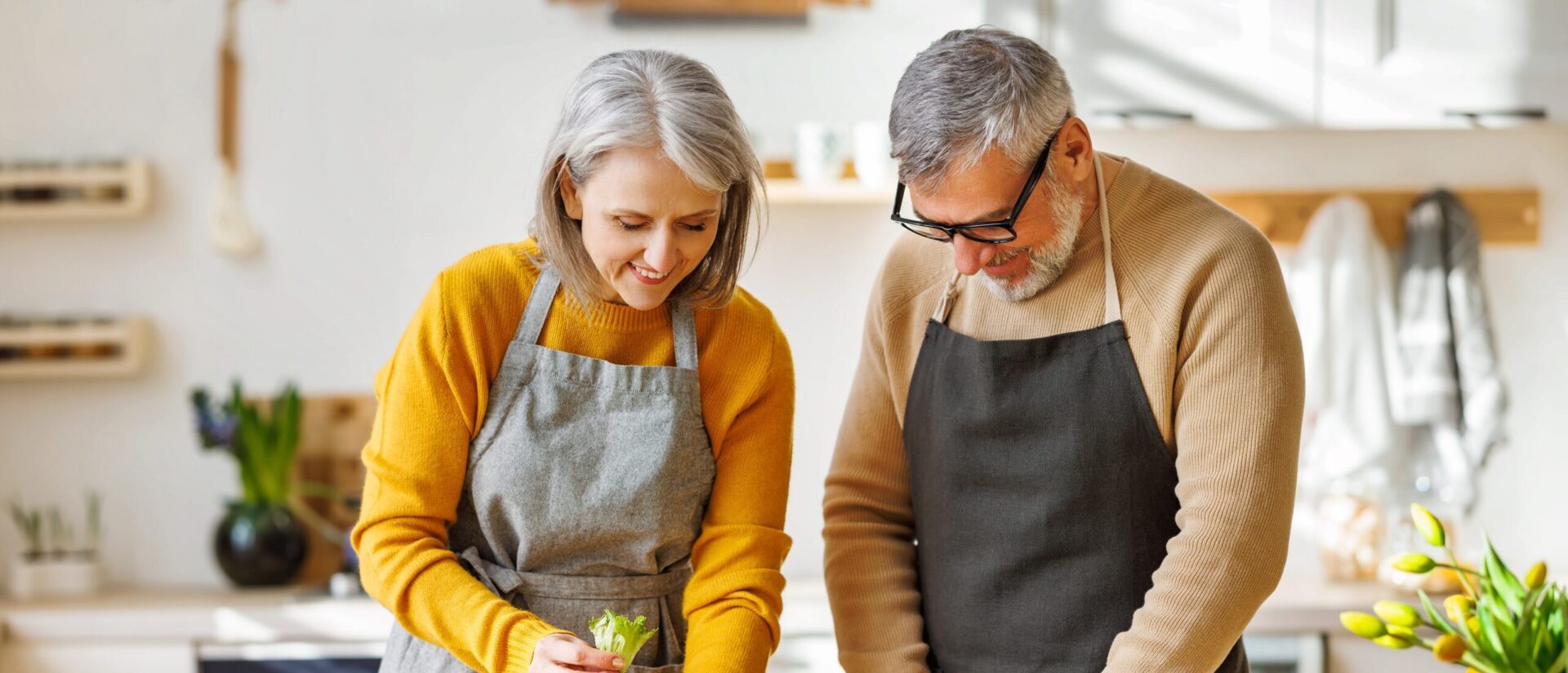 Two people wearing aprons are joyfully preparing food together in a bright kitchen, surrounded by plants and kitchen utensils.