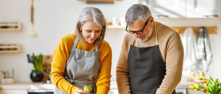 Two people wearing aprons are joyfully preparing food together in a bright kitchen, surrounded by plants and kitchen utensils.