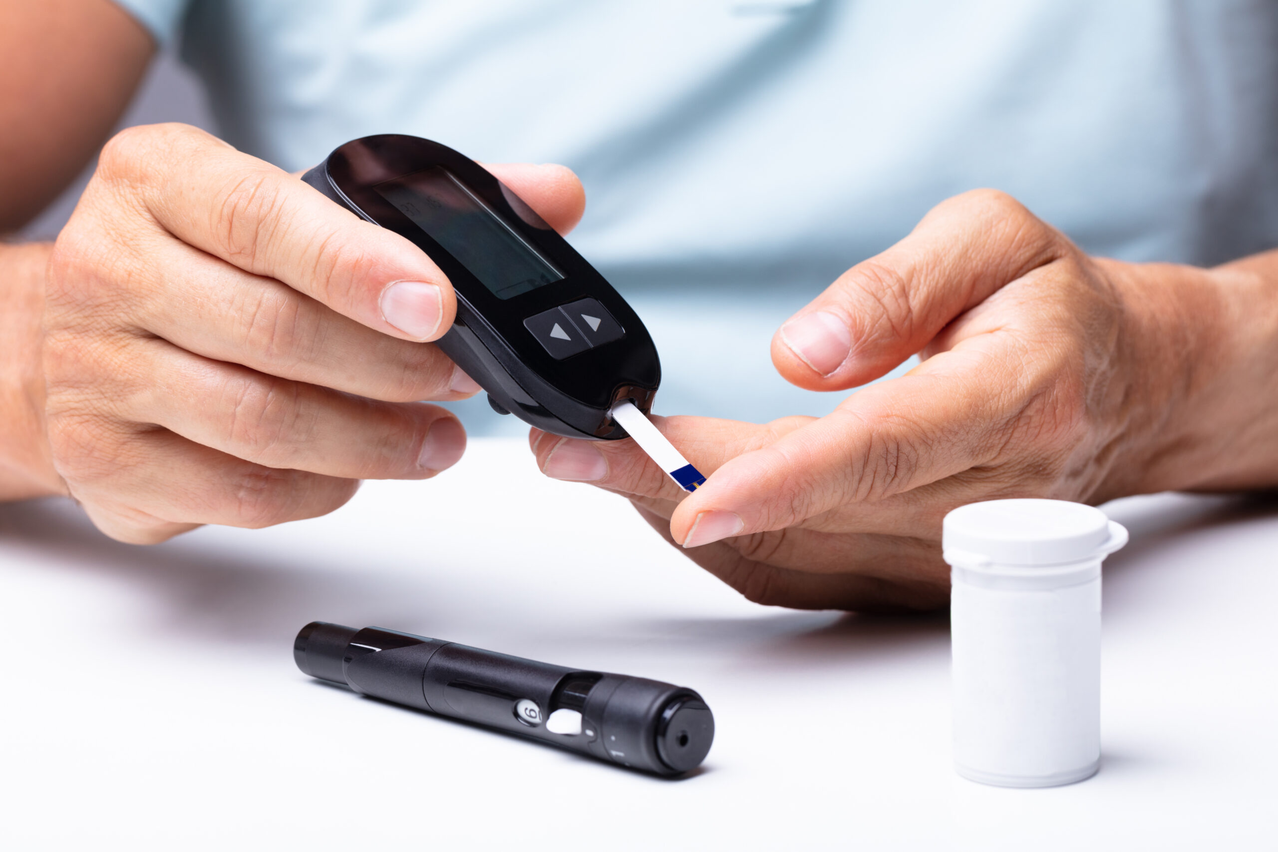 Hands hold a blood glucose meter and test strip, checking a fingertip; a white container and lancet device rest on a white table.