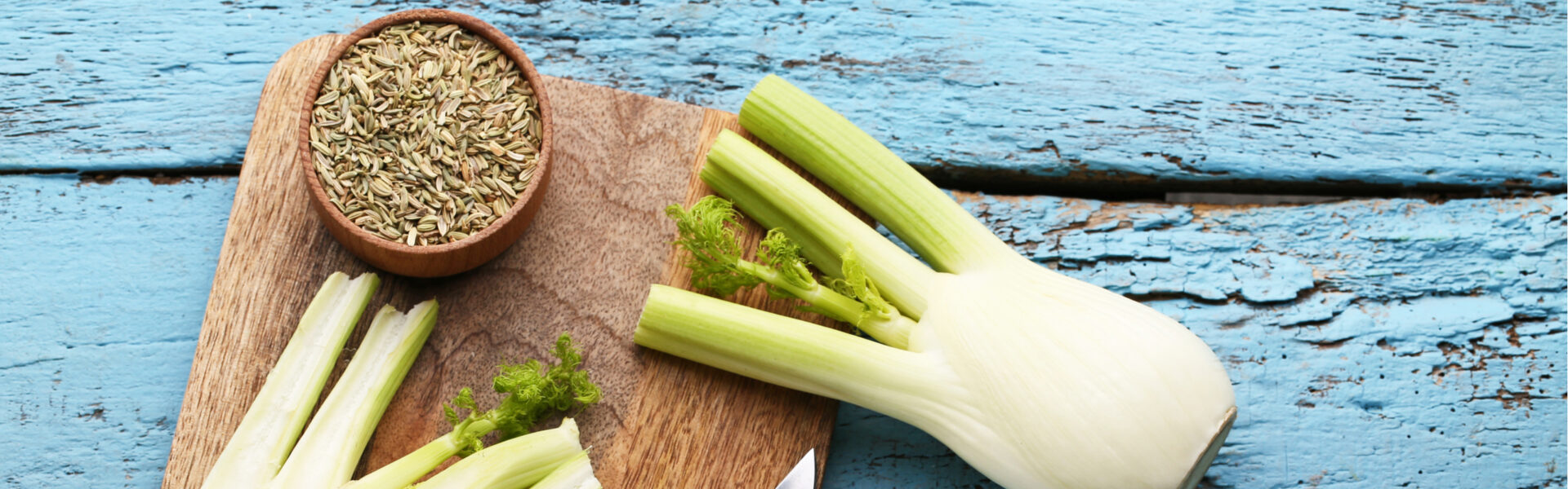 Fennel bulb and sliced stalks rest on a wooden cutting board beside a small bowl filled with fennel seeds, set against a textured, blue-painted wooden background.