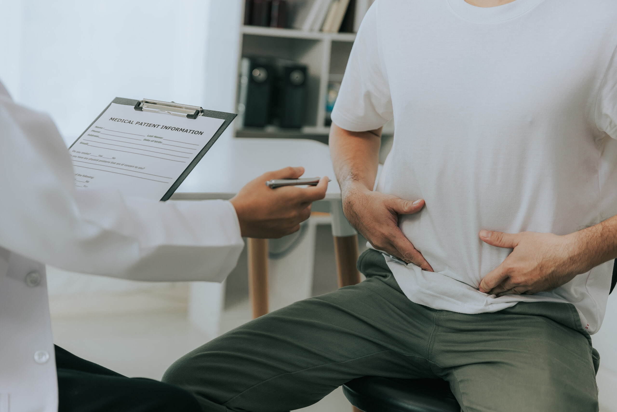 A doctor holds a clipboard labeled "MEDICAL PATIENT INFORMATION" while discussing with a patient who gestures towards their stomach, in a clinical office setting.