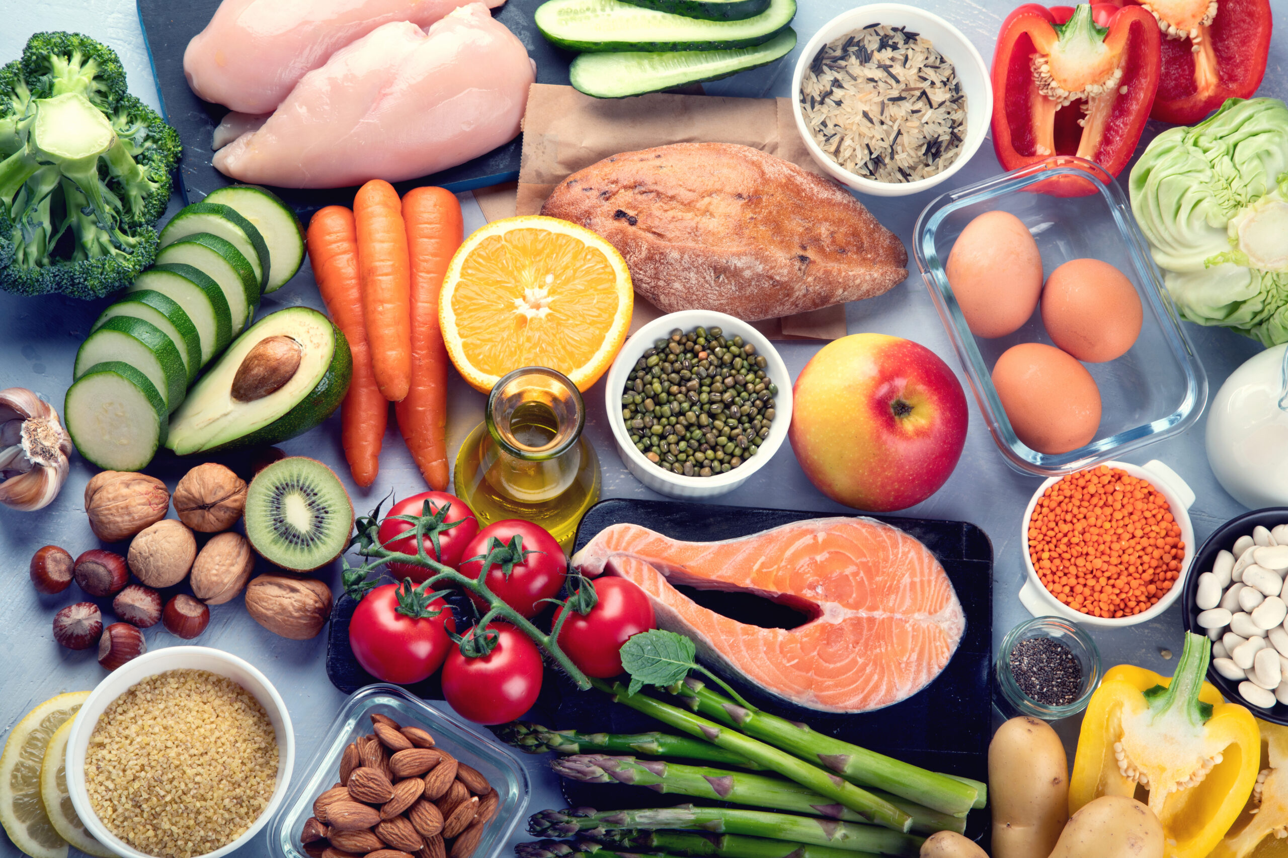 Various fresh foods lie arranged on a table, including salmon, chicken, vegetables, fruits, beans, nuts, grains, and olive oil, showcasing a diverse selection of healthy ingredients.