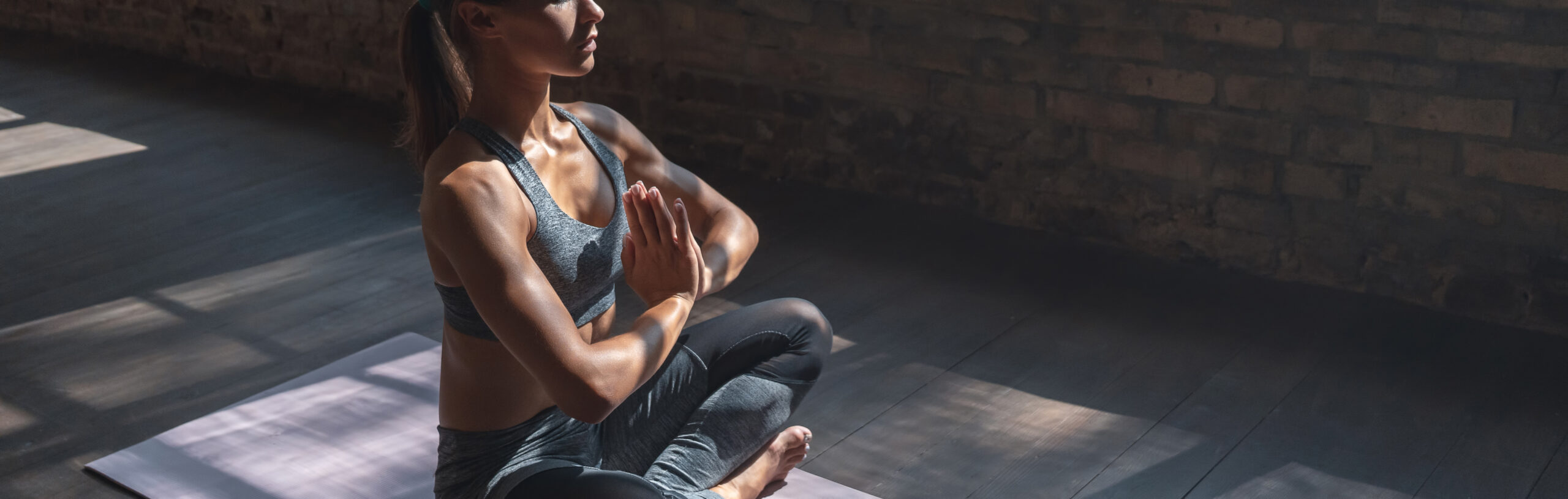 A woman sits cross-legged in a meditative pose with palms together, wearing athletic gear, on a yoga mat in a dimly lit room with wooden floors and brick walls.